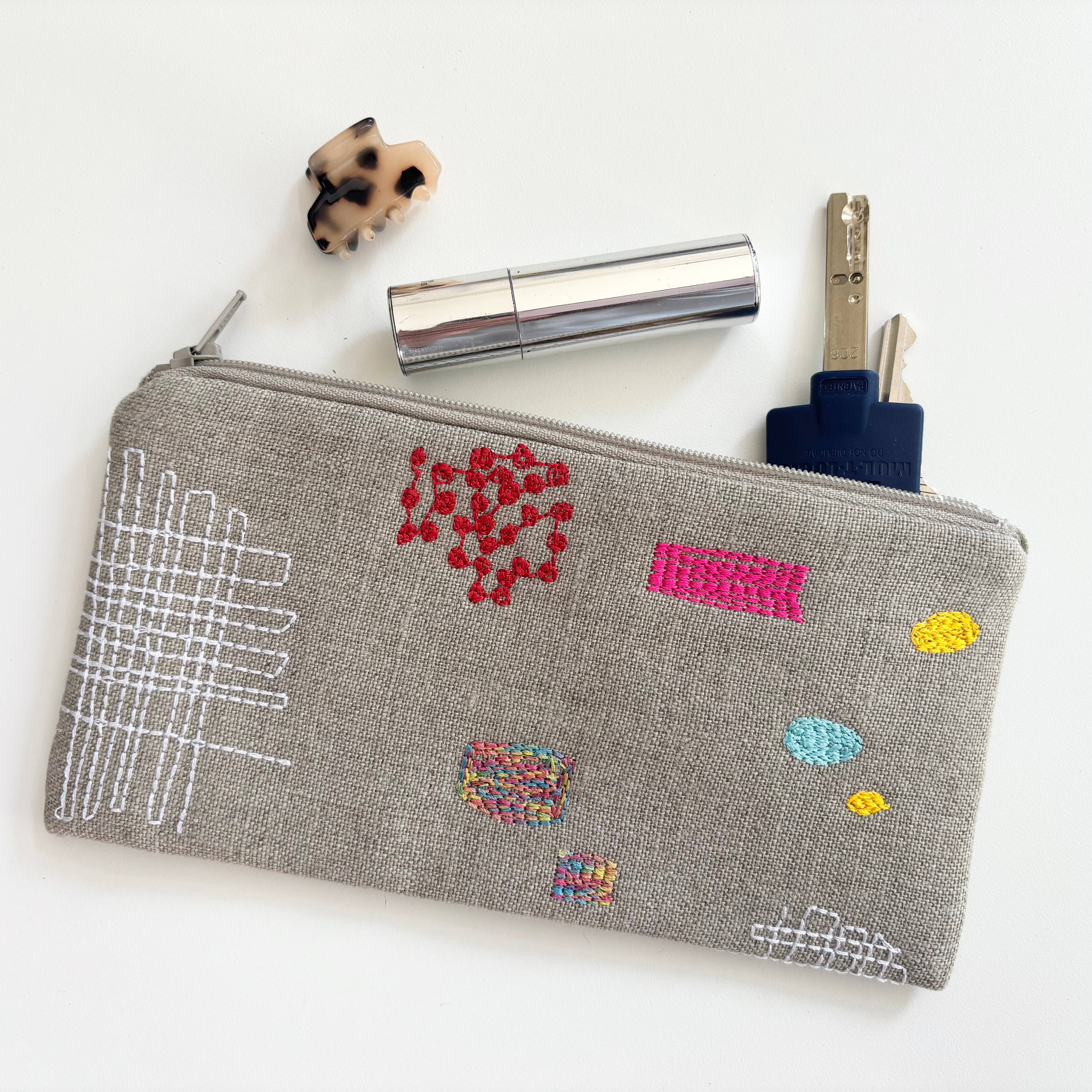 Patches Pouch by K Studio made of natural flax linen with colorful stitched patch motifs, slim rectangular shape, and zip closure on a neutral background.