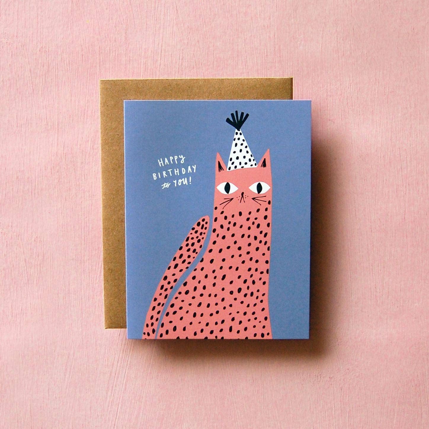 Spotted Cat Birthday Card by Badger & Burke featuring a pink spotted cat wearing a black-and-white party hat on a blue background.