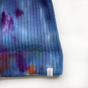 Painter's Hand-Dyed Beanie Hat by Merle Works in rich blue ribbed cotton-blend knit with hand-dyed splashes of purple, green, orange, and magenta, featuring a folded cuff and woven logo tag.