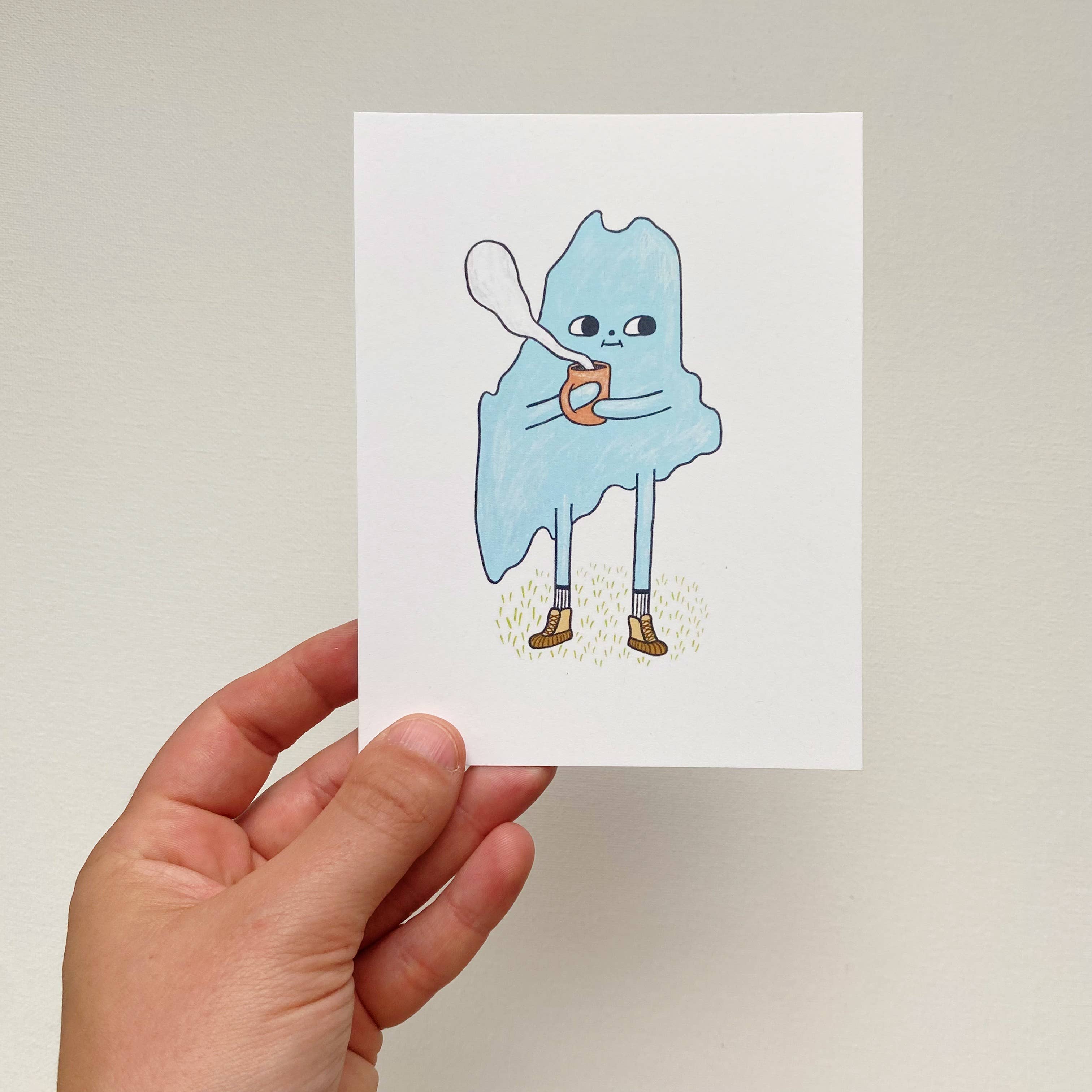 Cozy Maine Postcard by Allison McKeen featuring a blue illustrated character bundled up and holding a warm mug with “Maine” lettering.