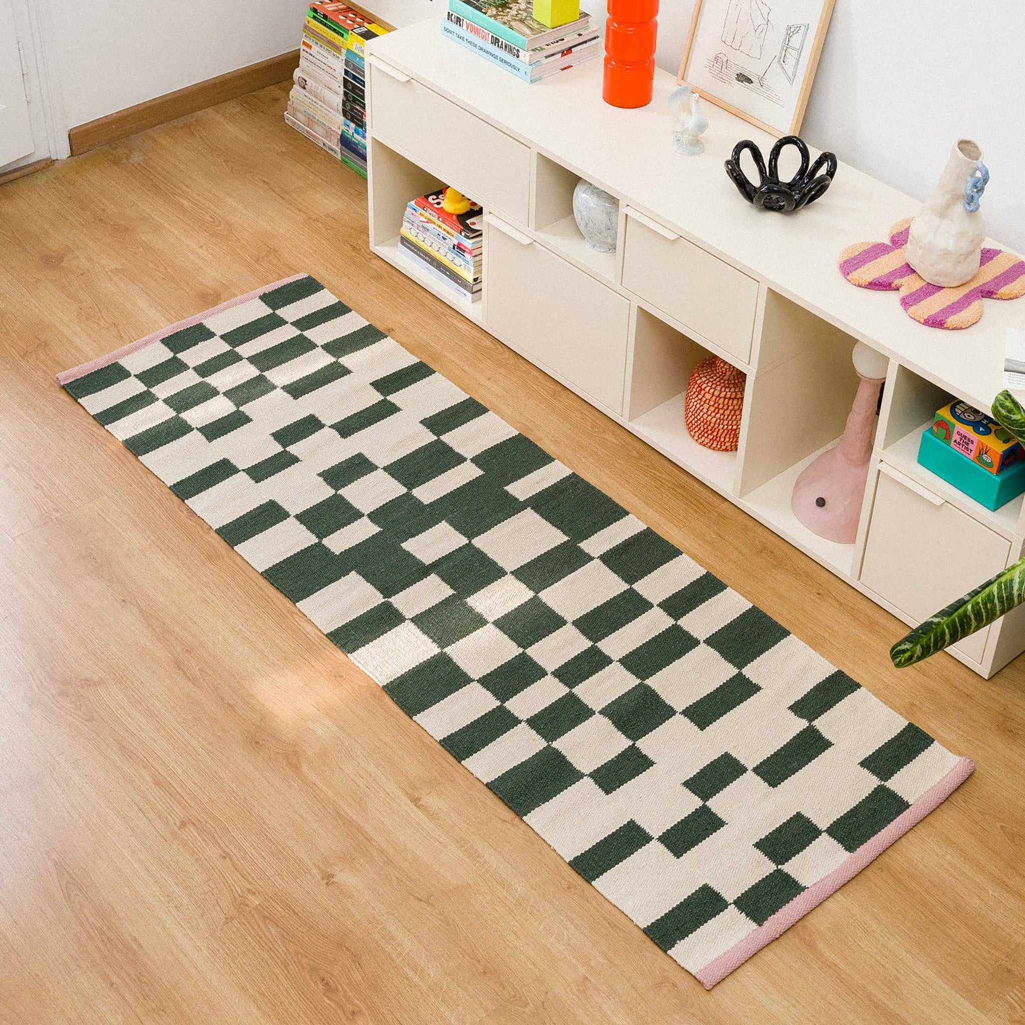 Blox Rug by Tarta Gelatina in Rosin, featuring an irregular forest green and cream checkerboard pattern with soft pink trim at both ends. Handmade from 100% cotton.